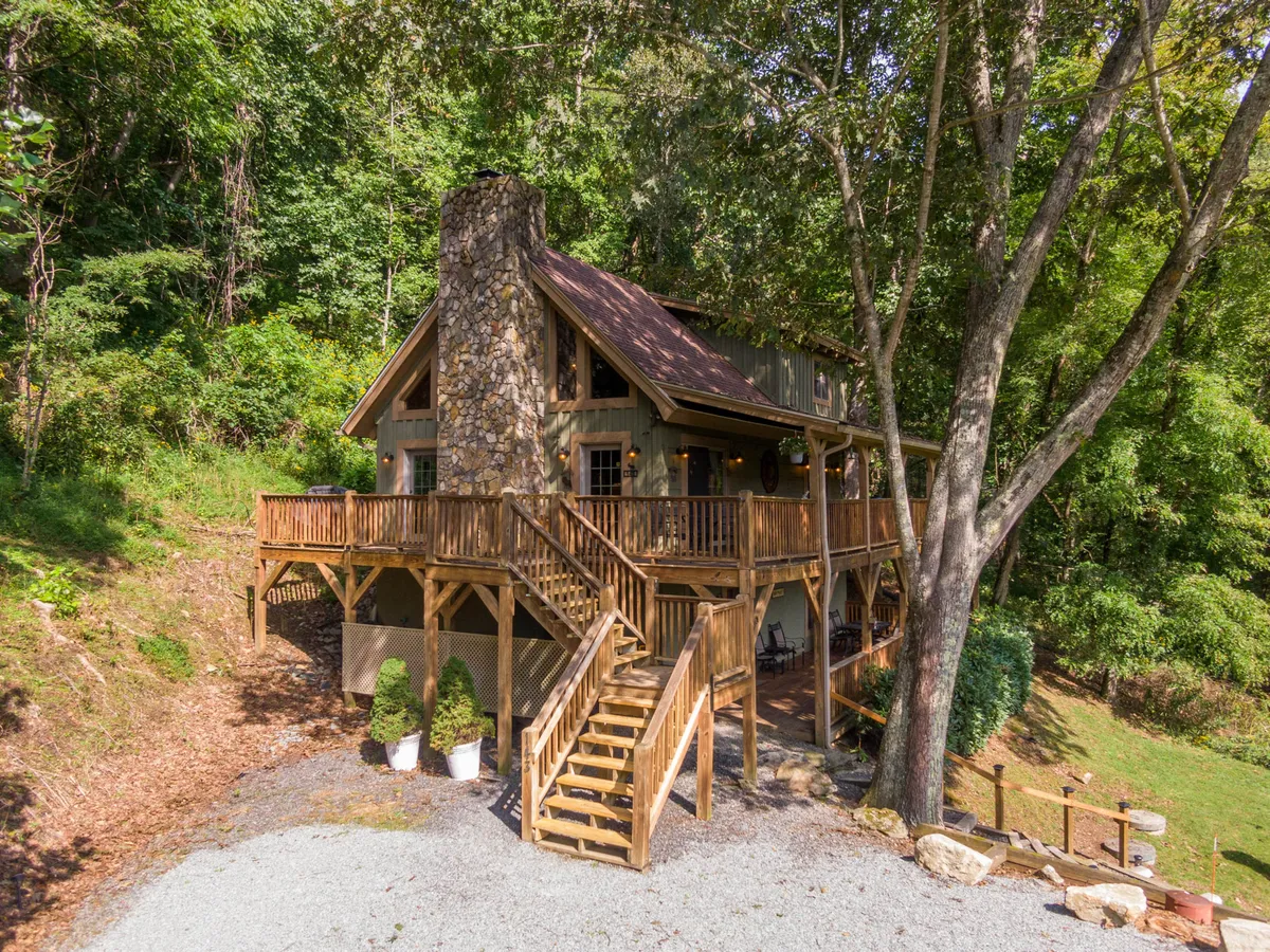 The image showcases the cabin's charming wooden deck surrounded by lush trees, with a striking stone chimney hinting at a cozy fireplace inside. The deck offers sweeping views ideal for sunset watching or stargazing, all set in the tranquil woods of Sugar Grove.