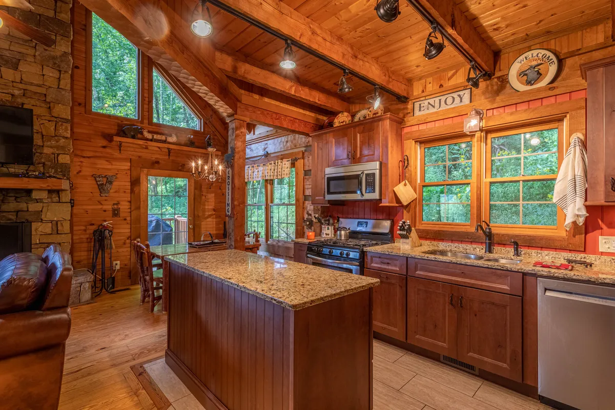 The image centers on the kitchen, featuring a striking granite countertop, wooden cabinetry, large windows, a chandelier, and a glimpse of the stone fireplace. The kitchen is clearly a lively gathering spot, perfect for preparing meals while staying connected to others in the great room.
