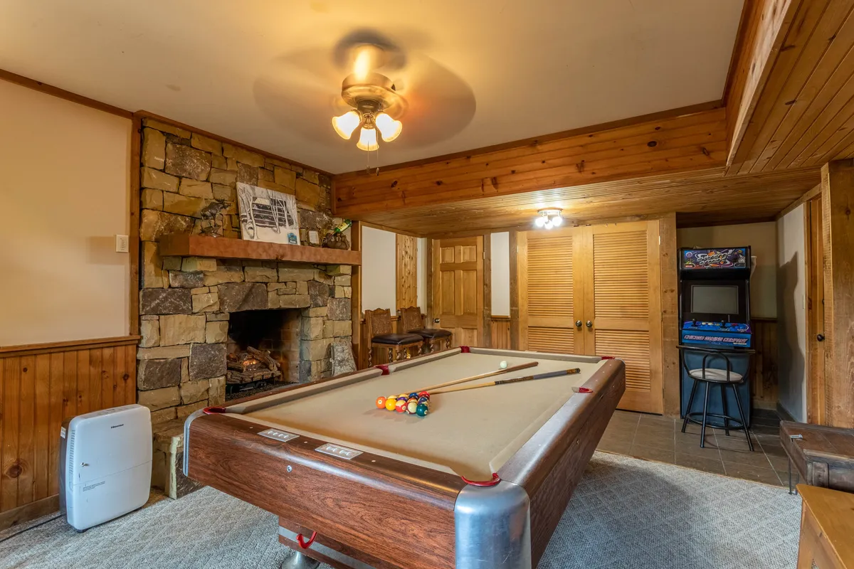A spacious game room featuring a regulation pool table as its centerpiece, set against a backdrop of warm wooden paneling. A rustic stone fireplace adds ambiance, while a ceiling fan and soft lighting create a relaxed, social atmosphere. The space is designed for group activities, with ample room for guests to gather, play, and converse.