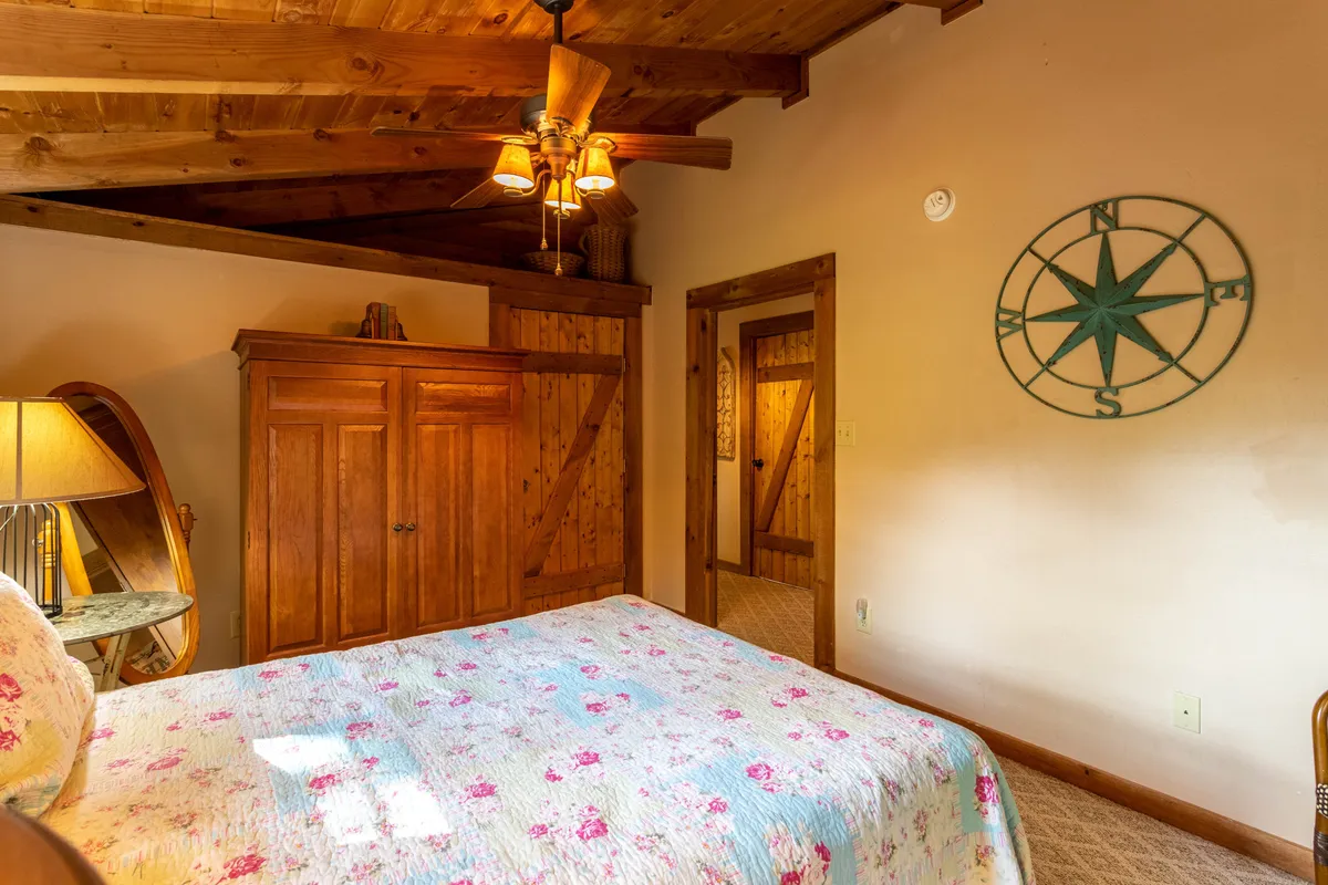 A bedroom with a quilted bedspread, compass wall decor, wooden ceiling beams, and a nightstand with a lamp, bathed in warm natural light. The scene feels peaceful and inviting, offering a sense of personal space and privacy.