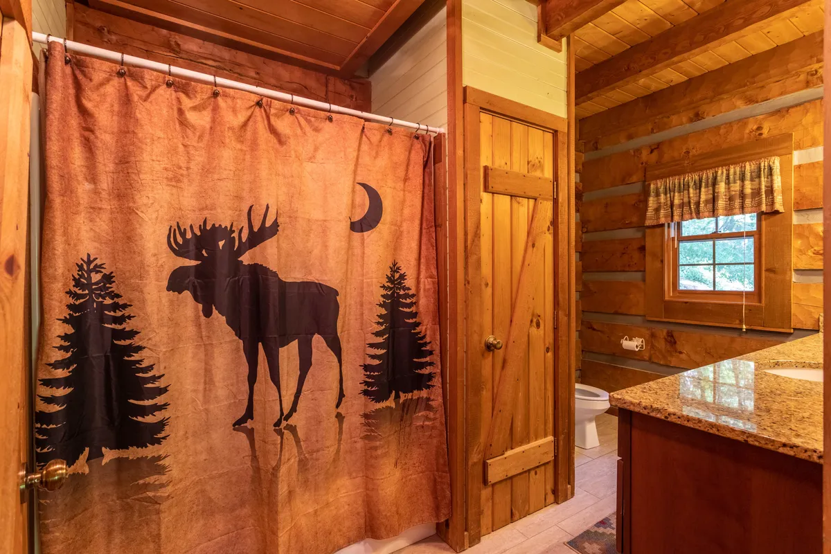 A tranquil bathroom scene featuring a moose-themed shower curtain, wooden paneling, and a granite countertop with a window view. The space feels rustic, clean, and serene—ideal for a relaxing bath or morning routine.