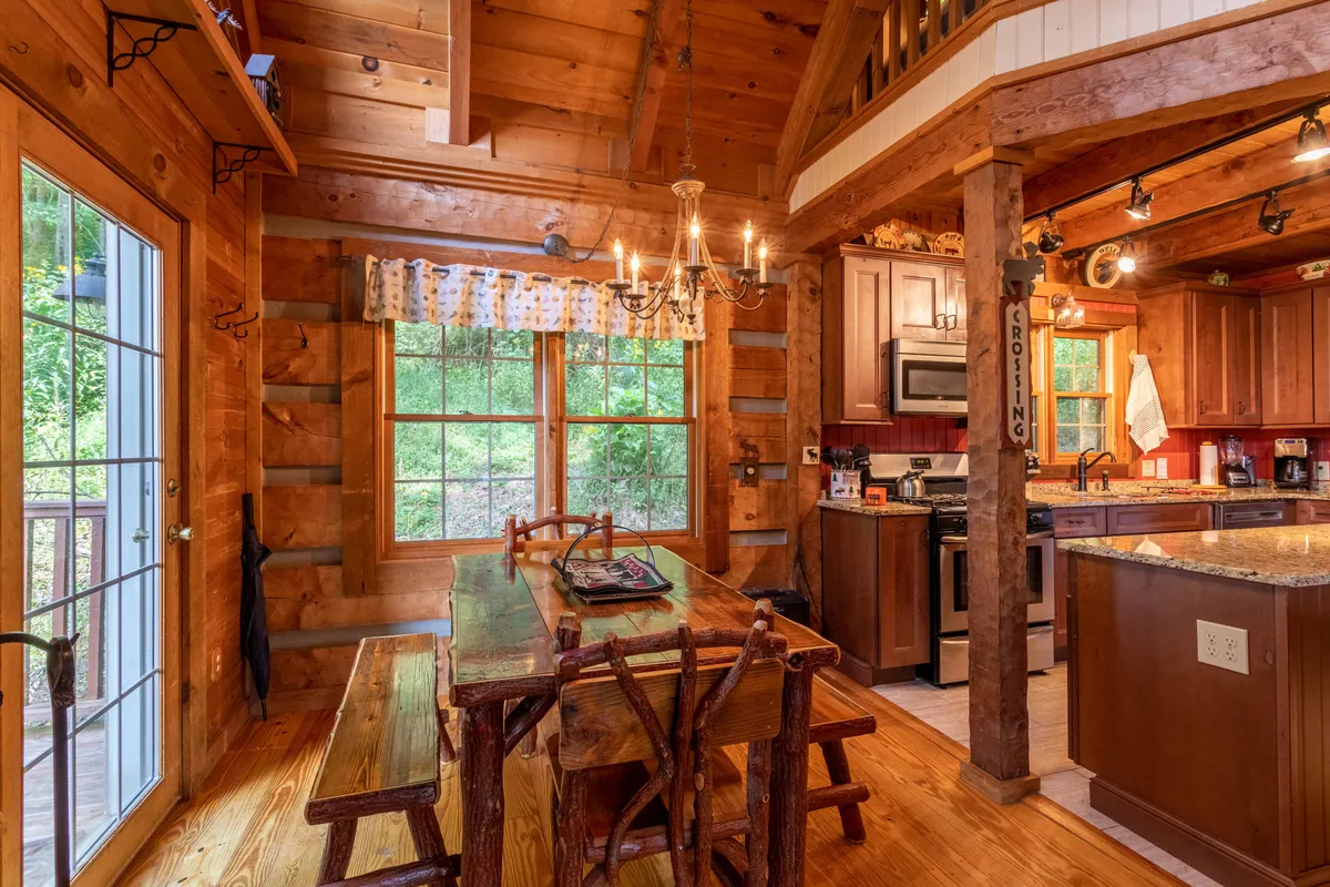 A warm dining area is shown here, with a sturdy wooden dining table beneath a chandelier, large windows letting in natural light, and the kitchen counter nearby. The scene is set for shared family meals and relaxed mornings.