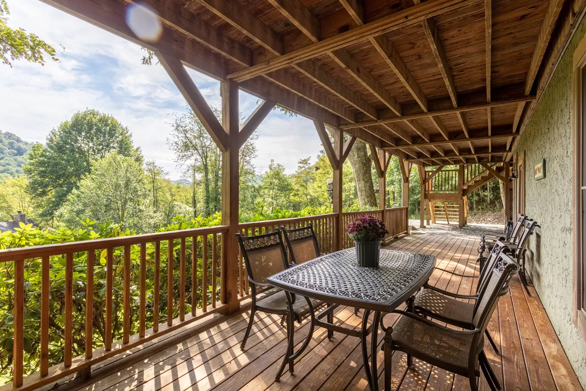 This exterior image features a wooden dining table and chairs on a deck surrounded by lush greenery, with exposed beams overhead. The setting invites groups to dine outdoors, relax, and socialize while immersed in nature. The expansive deck and serene environment directly connect to the mountain landscape, offering the perfect backdrop for group gatherings.
