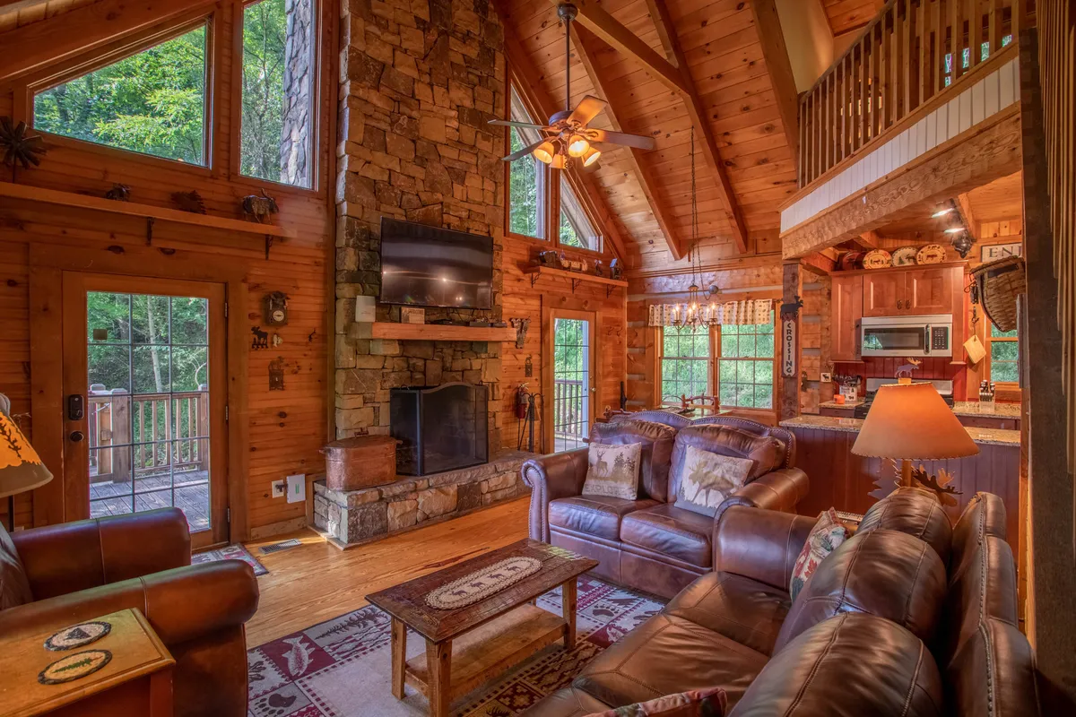 This image showcases the great room with a large stone fireplace as the focal point, flanked by leather sofas and expansive windows. Exposed wooden beams stretch overhead, creating a sense of openness and mountain character. The mood is warm and welcoming, with natural light illuminating the space and inviting relaxation or social gatherings.