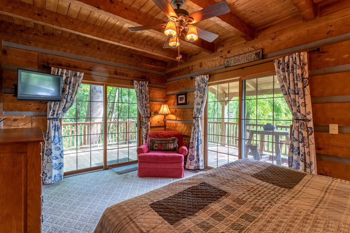 A comfortable pink chair faces large windows that offer a sweeping view of the surrounding trees, with wooden furniture and a TV mounted on the wall. The natural light streaming in suggests easy access to outdoor adventure and a connection to nature right from the main living area.