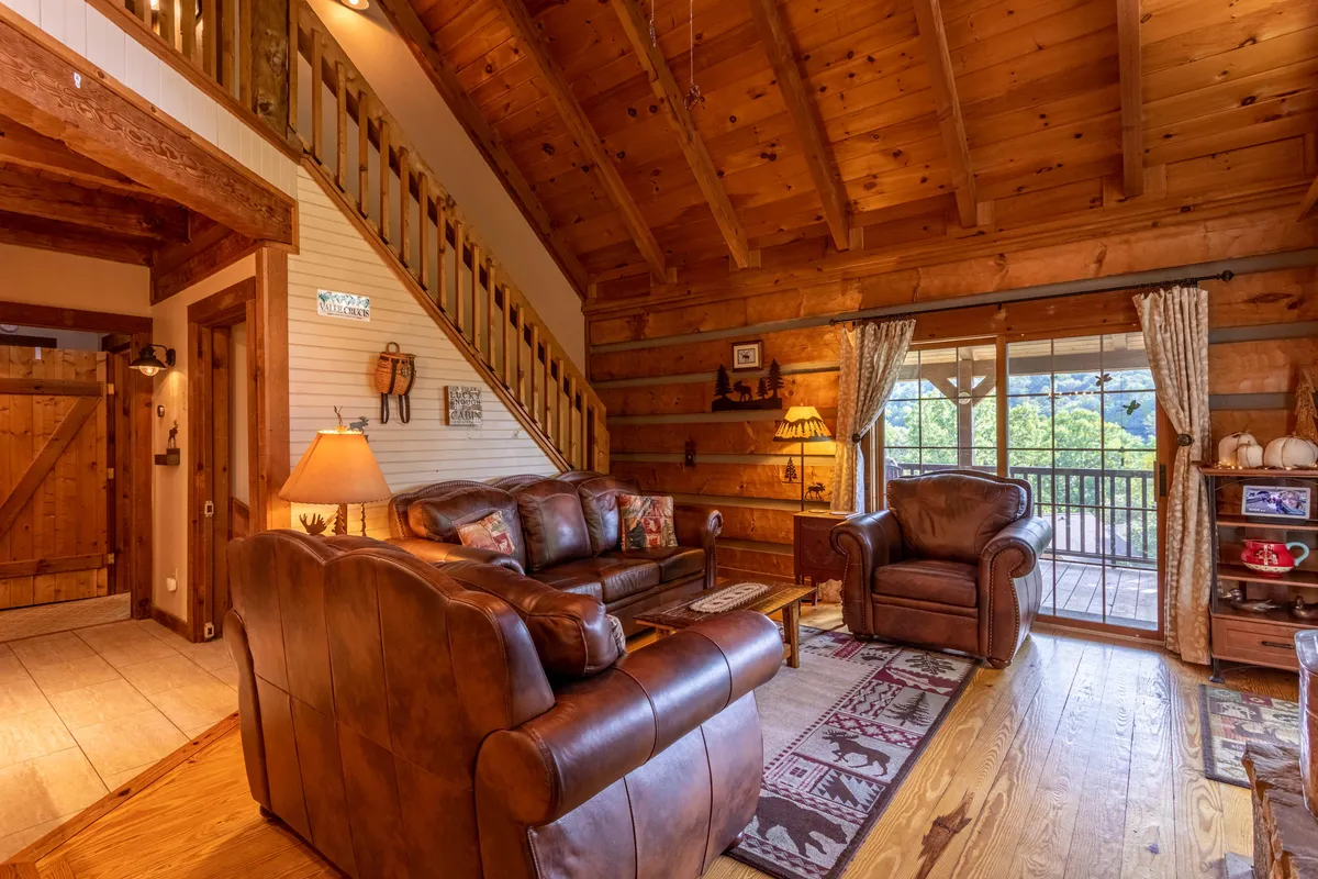 A cozy living area featuring leather sofas, a wooden staircase, and large windows that let in natural light. The space is arranged for relaxation, with the fireplace as a focal point, and decorative items on display that add charm and warmth. The views through the windows connect the interior to the scenic surroundings, suggesting a tranquil setting ideal for peaceful retreats.