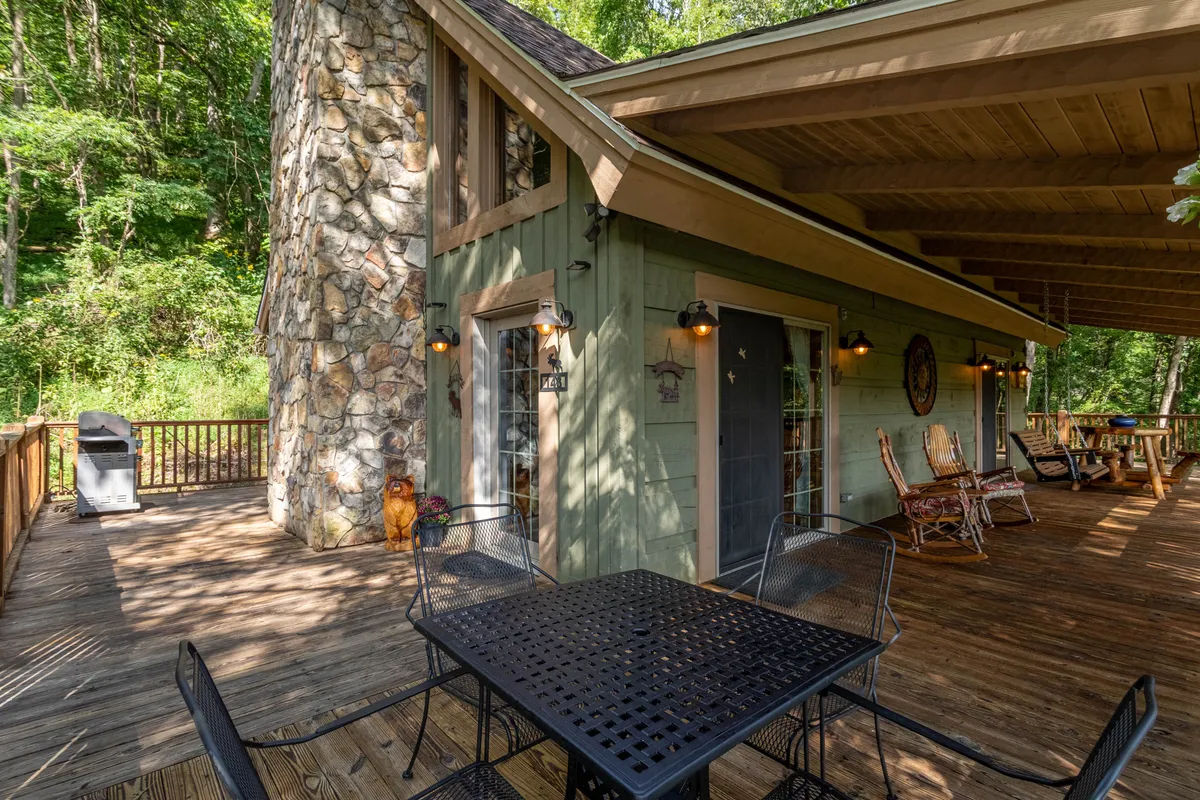 A wide view of the cabin's exterior with a prominent stone chimney, wooden deck, outdoor seating, and a green exterior wall. The deck faces surrounding trees, inviting guests to dine outside, barbecue, or simply bask in the serenity of the mountains.