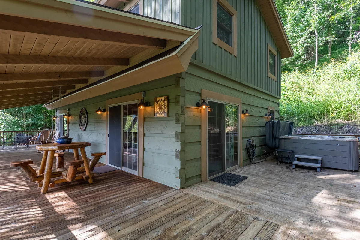 This image shows a spacious wooden deck with a wooden dining table, comfortable outdoor seating, a bubbling hot tub, and sliding glass doors leading into the home. The green siding of the house and surrounding trees enhance the sense of privacy and immersion in nature.